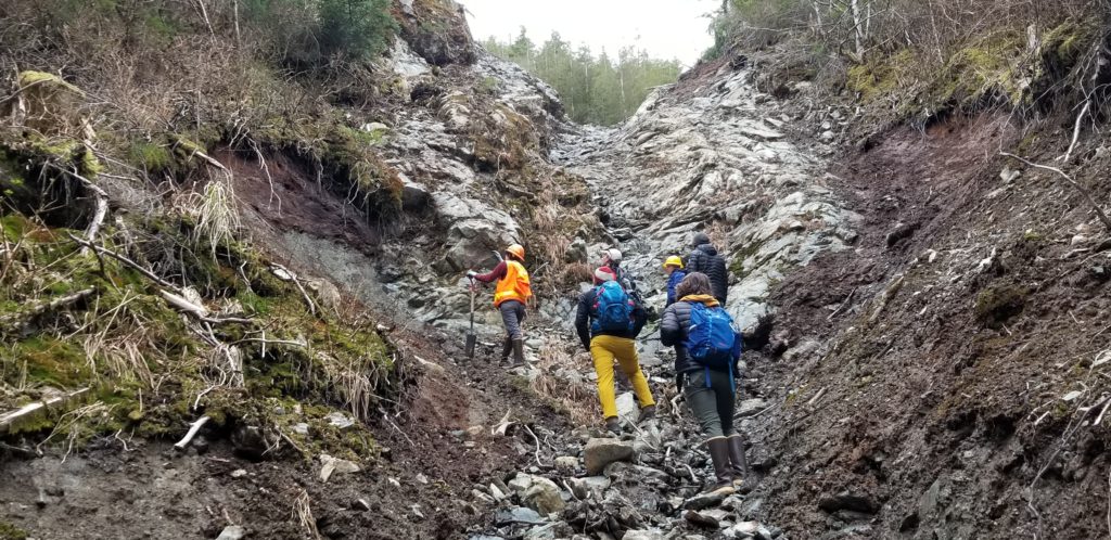 Six people standing on a steep hillslope where debris has slide down. Standing on gray rock next to brown soil. 